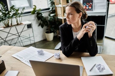 A middle-aged woman sits at a desk surrounded by crumpled paper, showing signs of stress.