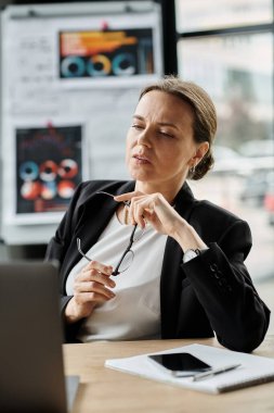 Middle-aged woman sitting at a desk, overwhelmed by stress while working on a laptop. clipart