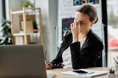 A middle-aged woman sits at a desk with a laptop, showing signs of stress and mental strain.