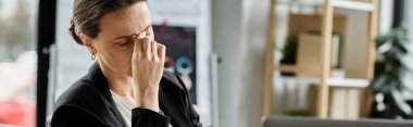 Middle-aged woman sits at desk, covering eyes, stressed.