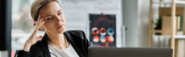 Middle-aged woman sits in front of laptop, hand on head, visibly stressed.