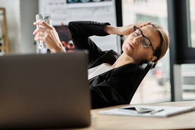 Woman holds water near laptop in stressful office environment.