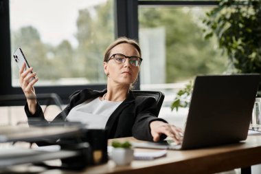 A middle-aged woman is engrossed in work at her desk with a laptop and cell phone.