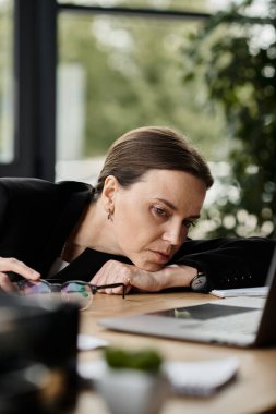 Middle-aged woman experiencing stress is hunched over her laptop in an office setting. clipart