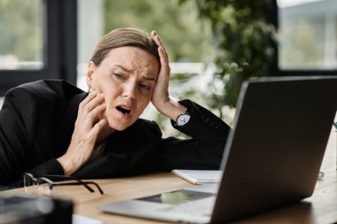 Woman in distress, holding head at cluttered desk.