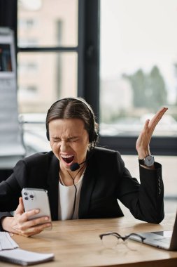 Middle-aged woman in distress shouting at smartphone while seated at desk. clipart