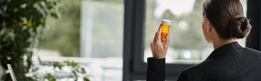 Woman seeks refuge by window, holding medicine bottle.