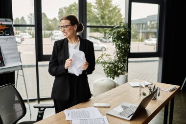 Middle-aged woman in business suit, looking stressed, standing in front of window. clipart