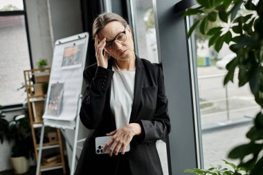 A middle-aged woman in a business suit stands before a plant, her demeanor reflecting stress and melancholy.