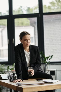 A middle-aged woman appears stressed, sitting at a desk with glass of water.