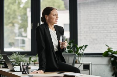 A middle-aged woman in distress sits at a desk, holding a glass of water.