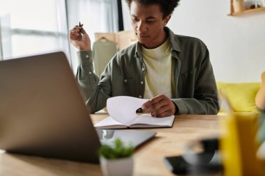 A young man focused on his laptop and notebook at a table, studying at home.