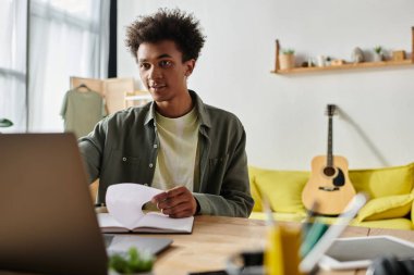 A young man, African American, sits at a desk with a laptop, engrossed in online studying.