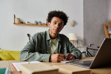 Young African American man focused on studying online using laptop and notebook at home.