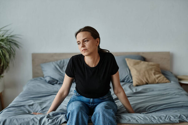 A middle-aged woman sits on top of a bed, lost in thought, amid a mental breakdown.