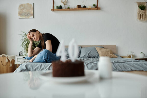 Middle-aged woman sitting with cake on bed, lost in thought.