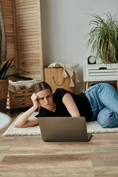 stock image A middle-aged woman lost in her laptop while laying on the floor at home.