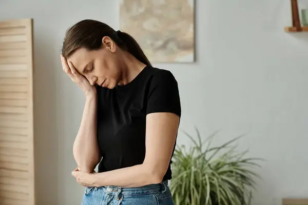 stock image Middle aged woman in distress holds head while standing in living room.