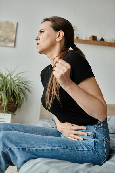Middle-aged woman experiences stomach discomfort while sitting on bed, struggling with depression.