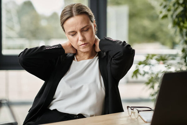 Middle-aged woman with neck pain, stressed, sitting in front of laptop.