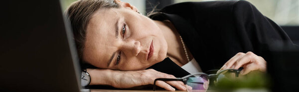 Middle-aged woman laying on desk, head on laptop, overwhelmed and stressed.