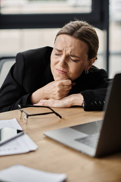 A middle-aged woman sits at a desk, hand on chin, deep in thought.