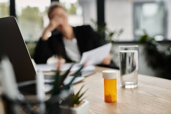 A woman, stressed and overwhelmed, sits at a desk with a bottle of medicine in front of her.