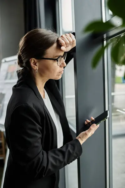 stock image A middle-aged business woman stands by a window, absorbed in her phone.