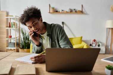 Young African American man talking on phone, working on laptop, and taking notes at desk.