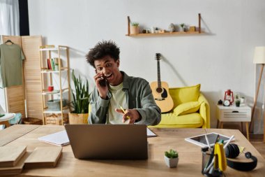 Young African American man talking on phone & using laptop at desk.