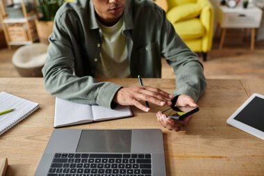 Young African American man studying online at home with laptop and cell phone.