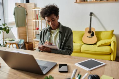A man sitting at a desk with a laptop open and a guitar beside him.