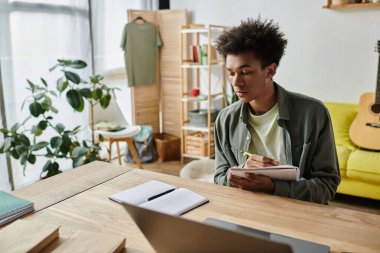 Young African American man studying at home, focused on writing in a notebook.