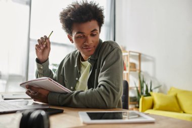 A young man, African American, studying at home, writing in a notebook.
