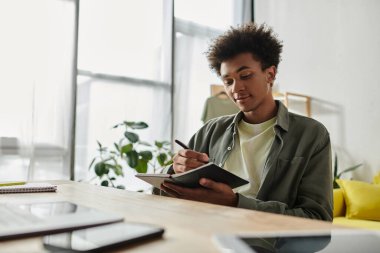Man of African descent sitting at desk, focused on writing in notebook.