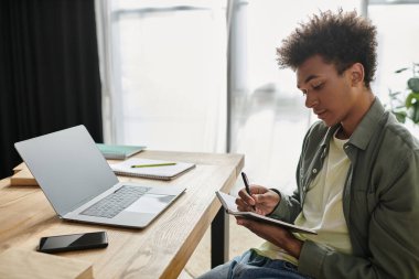 Young African American man focused on studying with notebook and laptop on desk.