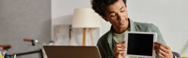 A man holds up a tablet in front of a laptop while studying online at home.