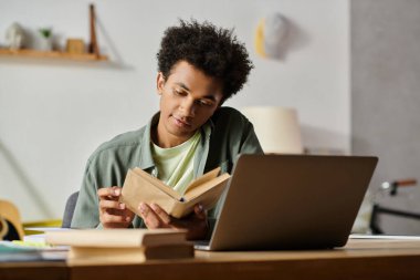 Young man immersed in book and laptop.