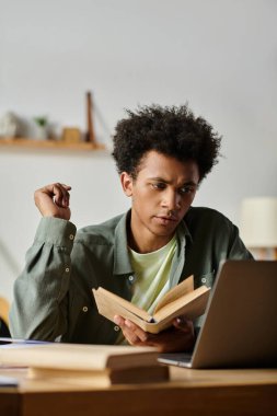 A young man engrossed in studying with a book and a laptop at a desk.