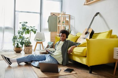 A young man of African American descent sits near a yellow couch with a laptop and guitar.