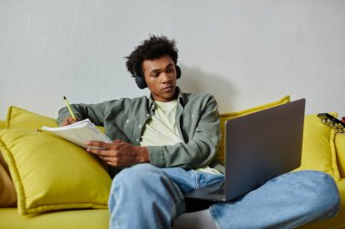 A young African American man studies online at home, sitting on a yellow couch with headphones and a laptop.