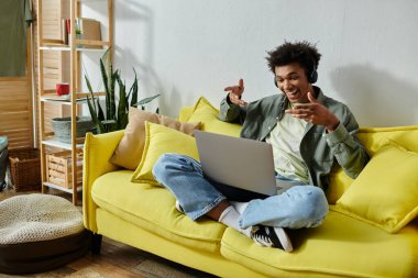 A young man engrossed in online studying, seated on a yellow couch with a laptop.