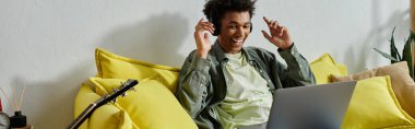 Young man, African American, sitting on yellow couch, using laptop for online study.