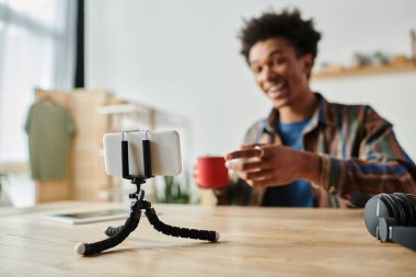 Young man sitting at table, recording video on phone tripod.