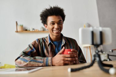 Young man, African American, smiles at camera while holding coffee cup. clipart