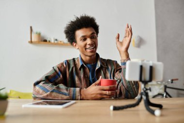 A young African American man sits at a table with a phone and a cup of coffee.