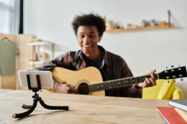 A young African American male blogger playing guitar on a tripod while speaking into a cell phone camera. clipart