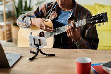 Young African American male blogger plays guitar while speaking on phone camera. clipart