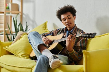 A young African American man playing an acoustic guitar on a yellow couch while talking on his phone. clipart