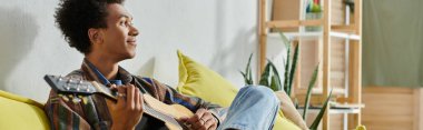A young African American man sits on a couch, playing an acoustic guitar.